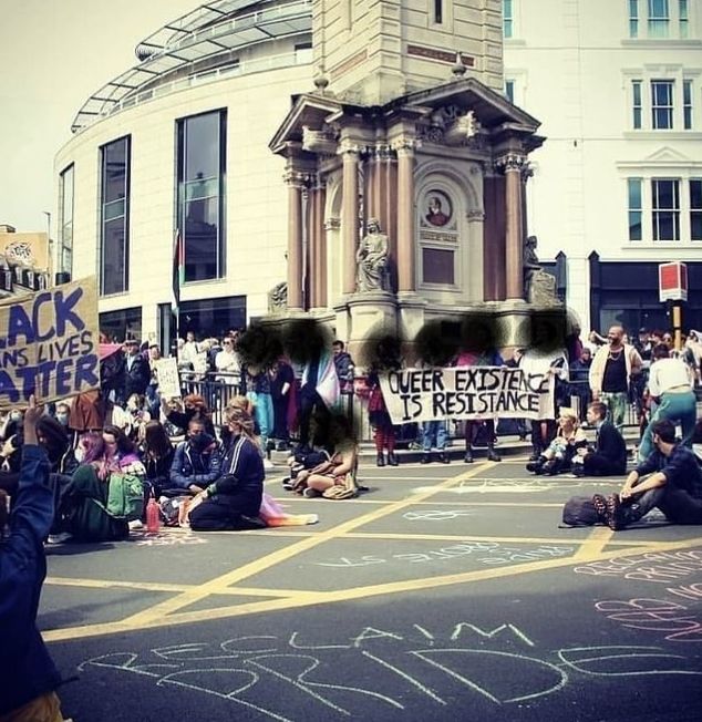 A crowd gathers around a monument for a Reclaim Pride event. Some protestors hold a banner that reads: 'Queer Existence is Resistance.'