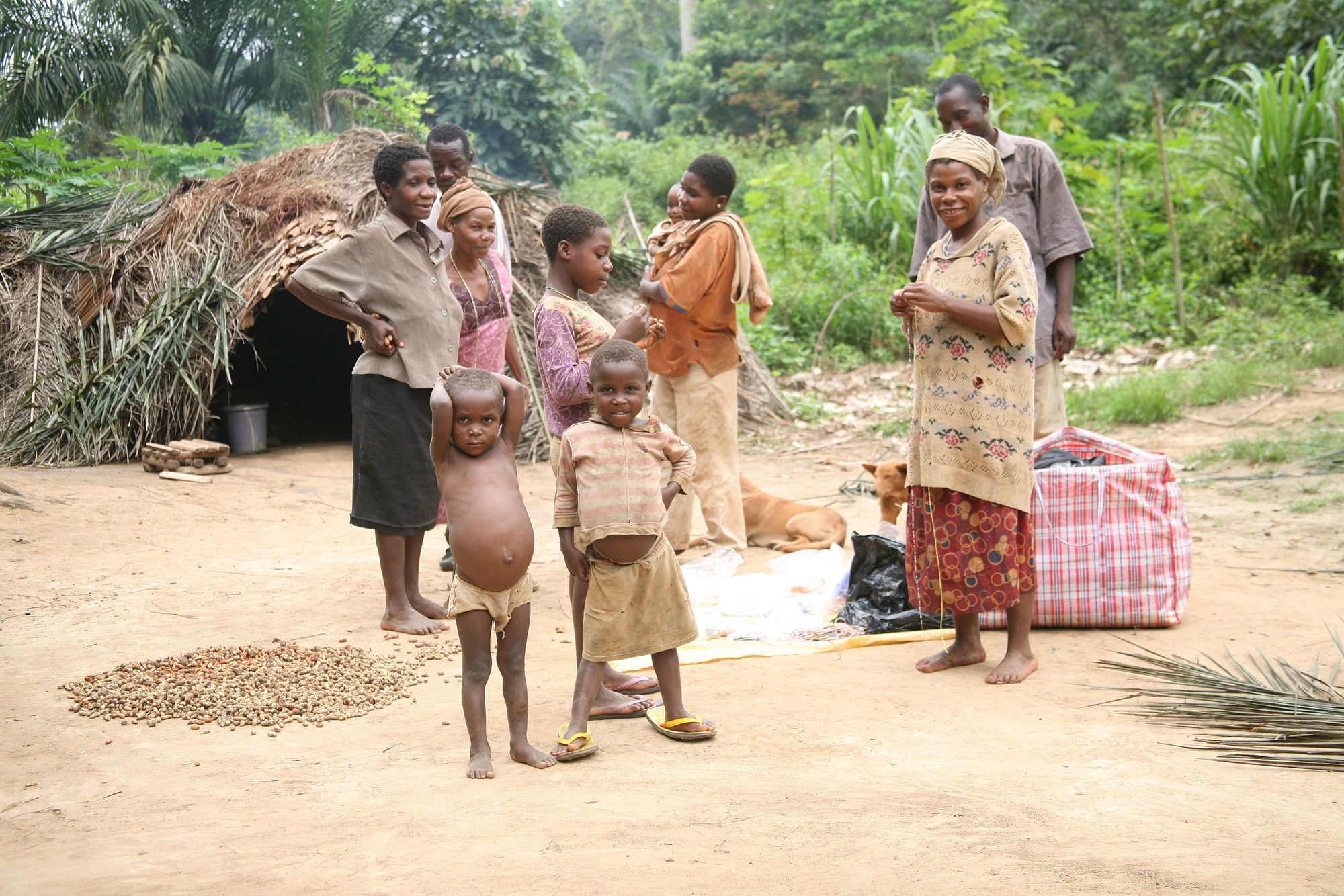 A group of Baka people standing in a rainforest village. In the foreground are two children, and the background a groups of adults. The scene depicts a mixture of modern garments and objects against a backdrop of a traditional hut constructed from local materials.