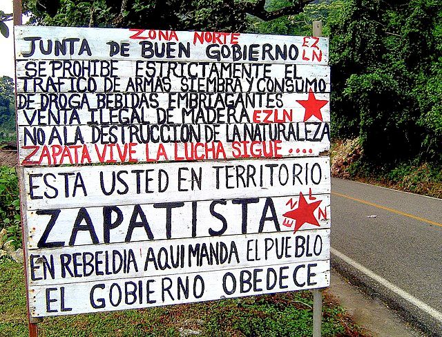Black and red text is painted on a white sign by the side of the road. There are EZLN star logos painted on the sign as well. Please see photo caption for English translation.