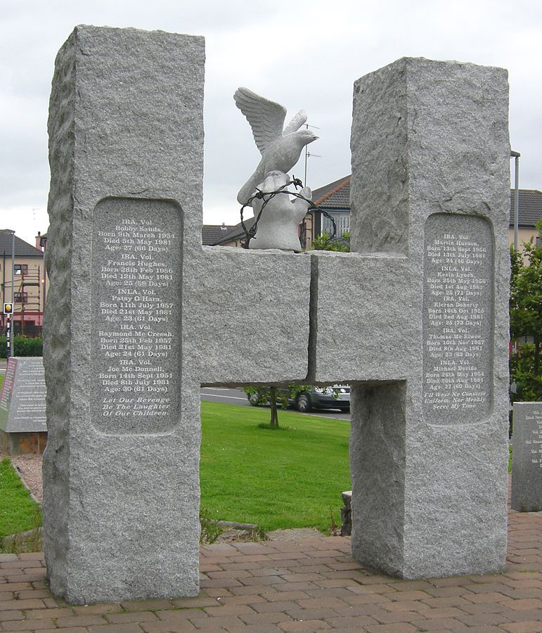 A h-shaped granite monument. Engraved are the names of the hunger striker. A hand sits atop the monument wrapped in barbed wire, with a dove perched on top.
