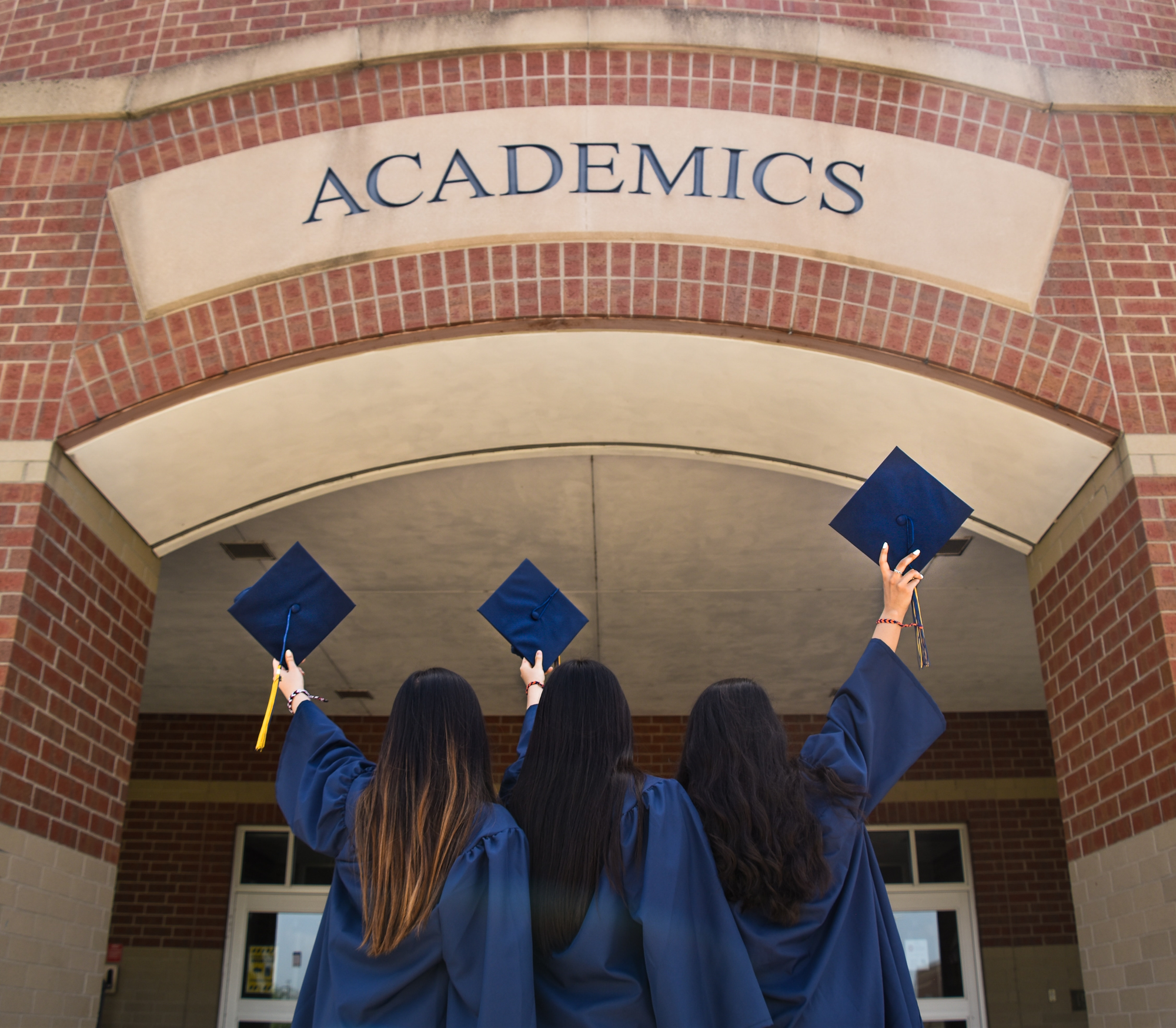 Three university students, in graduation gowns, holding their caps up and facing an academic building.