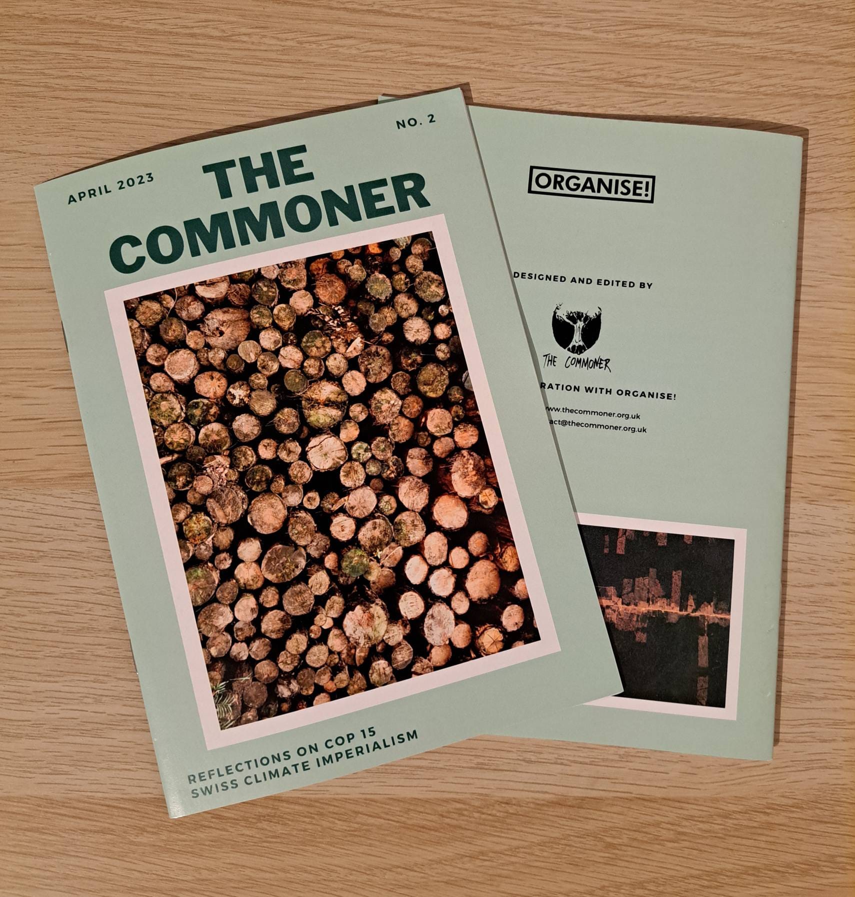 Two printed editions laid out on a wooden surface. The editions are green and contain a large frontcover image of logs. The edition is titled, 'The Commoner No.2.'